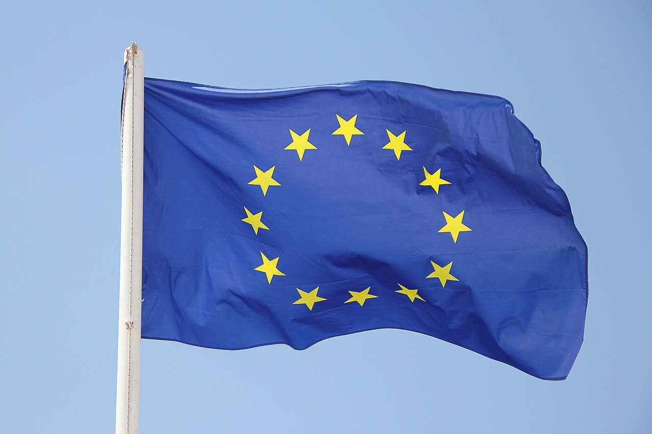 Die Flagge der Europäischen Union weht im Wind vor hellblauem Himmel. Die blaue Flagge zeigt den charakteristischen Kreis aus zwölf goldenen Sternen und ist an einem weißen Fahnenmast befestigt.