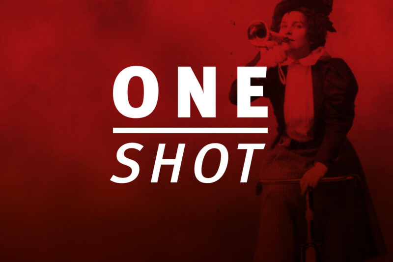 Logo 'ONE SHOT' in weißer Schrift auf rotem Hintergrund. Im Hintergrund ist schemenhaft eine Tänzerin in Bewegung zu erkennen. 'ONE' steht in Großbuchstaben über einem horizontalen Strich, darunter 'SHOT' in kursiver Schrift.