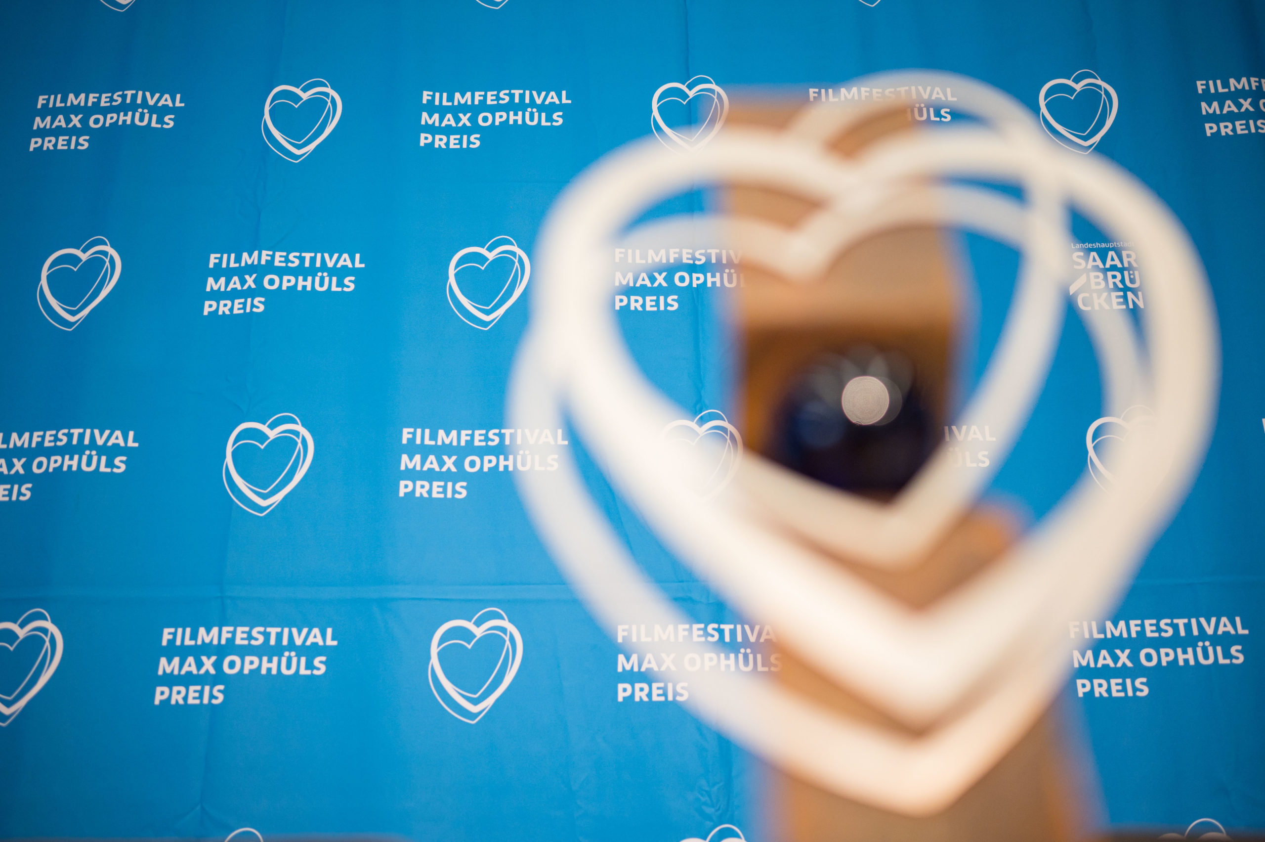 Eine unscharfe Nahaufnahme der Herz-Trophäe des Filmfestivals Max Ophüls Preis vor einem blauen Hintergrund mit dem Festival-Logo, bestehend aus weißen Herzen und Schriftzügen.