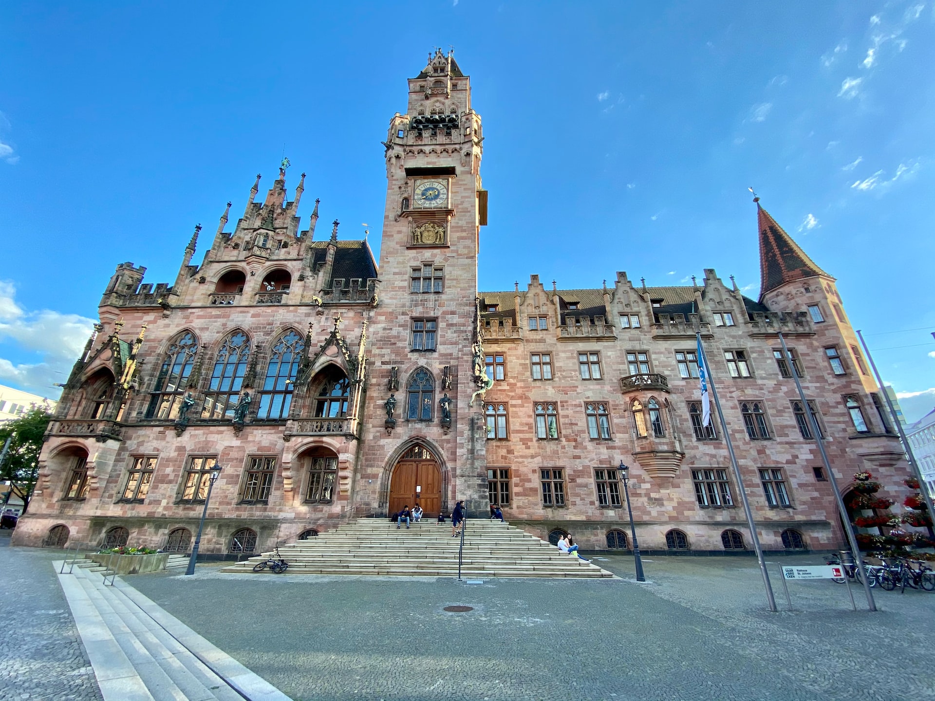 Historisches Rathausgebäude von Saarbrücken mit aufwendiger neugotischer Fassade und hohem Uhrturm, aufgenommen aus der Froschperspektive unter blauem Himmel; davor führen breite Stufen zum Eingang, auf denen einige Menschen sitzen.