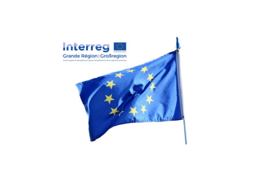 Eine EU-Flagge mit blauem Hintergrund und gelben Sternen weht an einem Fahnenmast. Über der Flagge befindet sich das Logo von Interreg Grande Région / Großregion, das das EU-Emblem und Text enthält. Das Bild steht für die Zusammenarbeit innerhalb der Europäischen Union, speziell im Rahmen des Interreg-Programms für die Großregion.