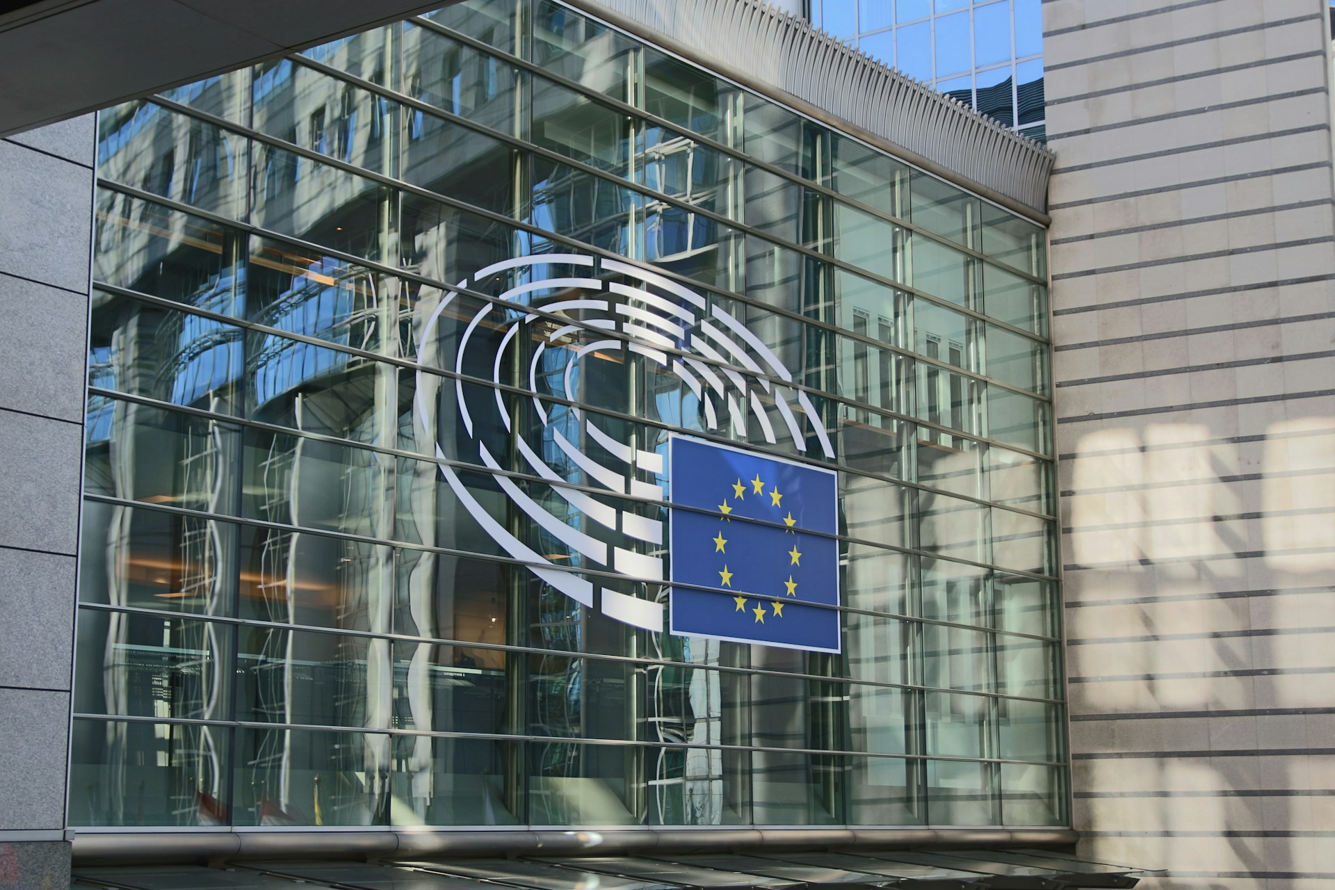 Eine Glasfassade eines modernen Gebäudes mit dem Logo des Europäischen Parlaments und der EU-Flagge. Die Reflexionen der Umgebung sind auf den Glasfenstern zu sehen.