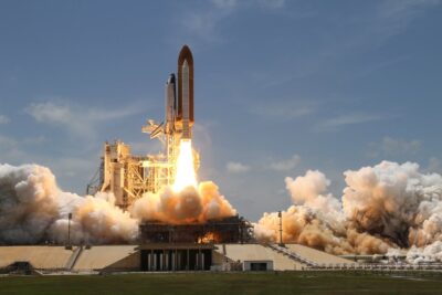 Ein Space Shuttle startet von einer Rampe in den Weltraum. Das Bild zeigt den Moment des Abhebens, mit dichten Rauch- und Feuersäulen, die aus den Triebwerken strömen. Im Hintergrund ist ein klarer blauer Himmel zu sehen.