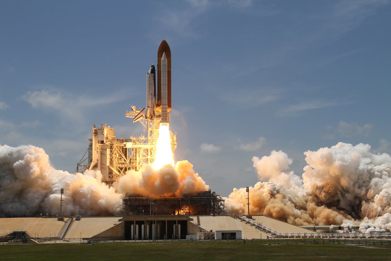 Ein Space Shuttle startet von einer Rampe in den Weltraum. Das Bild zeigt den Moment des Abhebens, mit dichten Rauch- und Feuersäulen, die aus den Triebwerken strömen. Im Hintergrund ist ein klarer blauer Himmel zu sehen.