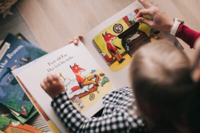 Eine Person hält ein Bilderbuch in den Händen, während ein kleines Kind auf ihrem Schoß sitzt und die Seiten betrachtet. Die Buchseite zeigt eine Illustration eines Fuchses in einem Schlafzimmer mit der Textzeile: „Poor old Fox. Has lost his socks.“ Im Hintergrund liegen weitere Kinderbücher verstreut. Die Szene vermittelt eine gemütliche Atmosphäre beim gemeinsamen Lesen.