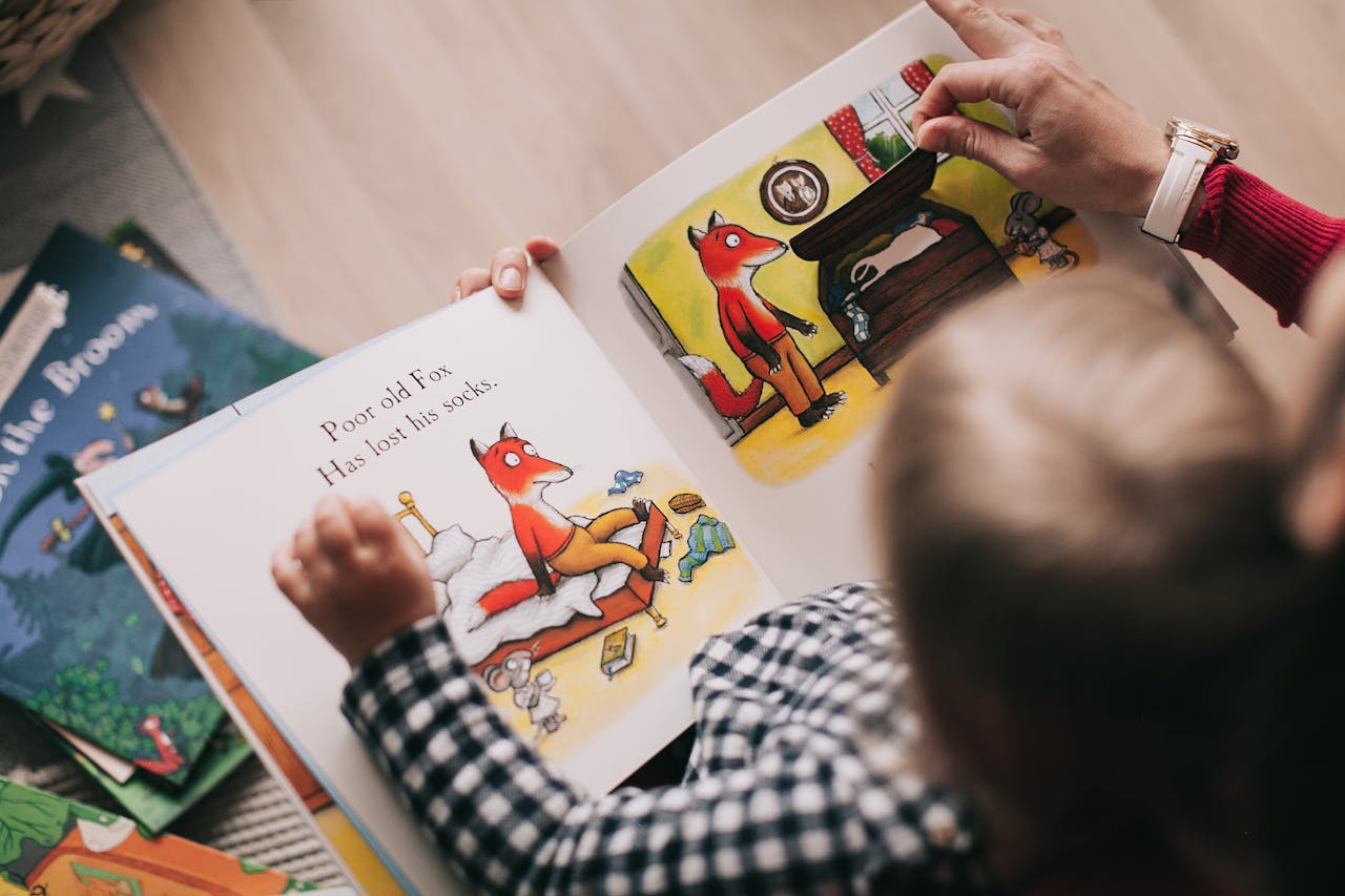 Eine Person hält ein Bilderbuch in den Händen, während ein kleines Kind auf ihrem Schoß sitzt und die Seiten betrachtet. Die Buchseite zeigt eine Illustration eines Fuchses in einem Schlafzimmer mit der Textzeile: „Poor old Fox. Has lost his socks.“ Im Hintergrund liegen weitere Kinderbücher verstreut. Die Szene vermittelt eine gemütliche Atmosphäre beim gemeinsamen Lesen.