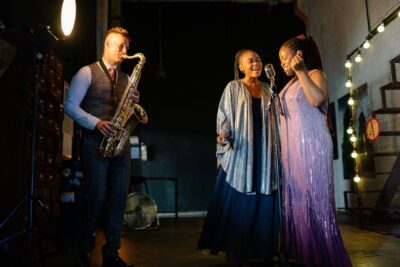 Drei Musiker:innen stehen auf einer Bühne. Links spielt ein Mann im Anzug Saxophon, während zwei Frauen in glitzernden, eleganten Kleidern rechts neben ihm singen. Die Bühne ist stimmungsvoll beleuchtet, und im Hintergrund sind Lichterketten zu sehen, die für eine warme Atmosphäre sorgen.