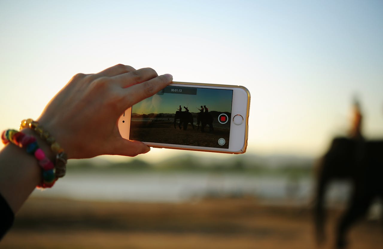 Ein Smartphone wird von einer Person mit bunten Armbändern gehalten, während ein Video aufgenommen wird. Im Display des Smartphones sind Silhouetten von Menschen zu sehen, die auf Elefanten reiten, und im Hintergrund sind Berge und ein Sonnenuntergang sichtbar.