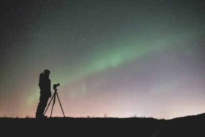 Die Grafik zeigt eine Person, die in einer dunklen, offenen Landschaft vor einem Stativ mit Kamera steht und in den Nachthimmel fotografiert. Der Himmel ist mit zahlreichen Sternen bedeckt, und es sind grünliche Lichter zu sehen, die möglicherweise das Nordlicht darstellen. Die Szene ist ruhig und vermittelt eine Atmosphäre der Stille und Naturverbundenheit, während die Person sich auf das Fotografieren des Himmels konzentriert.