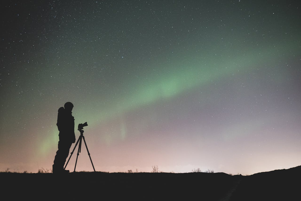 Die Grafik zeigt eine Person, die in einer dunklen, offenen Landschaft vor einem Stativ mit Kamera steht und in den Nachthimmel fotografiert. Der Himmel ist mit zahlreichen Sternen bedeckt, und es sind grünliche Lichter zu sehen, die möglicherweise das Nordlicht darstellen. Die Szene ist ruhig und vermittelt eine Atmosphäre der Stille und Naturverbundenheit, während die Person sich auf das Fotografieren des Himmels konzentriert.