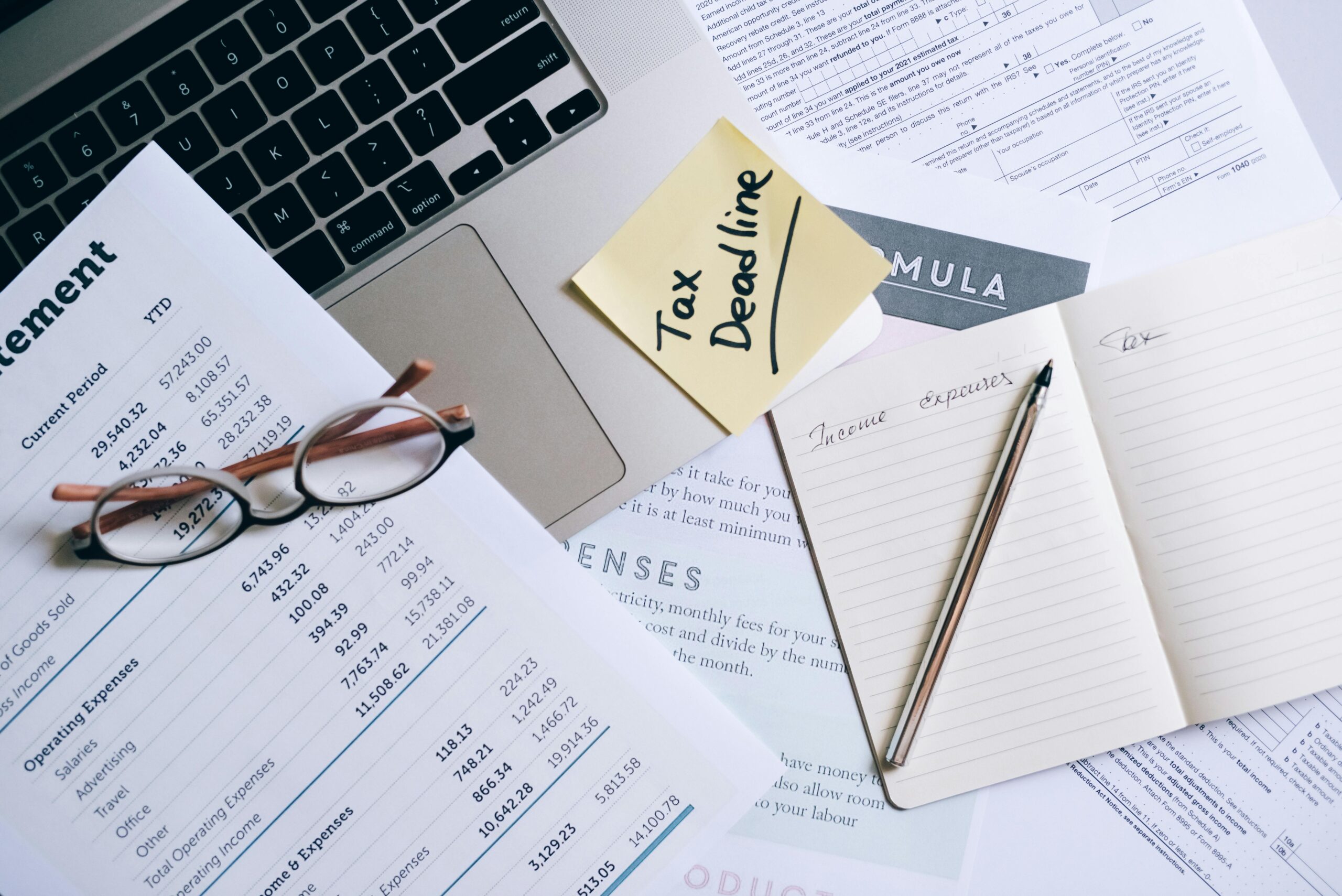 Das Bild zeigt eine Arbeitsfläche mit einem Laptop, mehreren Finanzdokumenten und einer Brille. Auf einem der Dokumente steht »Statement«. Ein Post-it mit der Aufschrift »Tax Deadline« liegt auf der Laptop-Tastatur. Daneben befindet sich ein offenes Notizbuch mit der Überschrift »Income & Expenses« und einem Stift darauf. Das Bild vermittelt das Thema Steuern und Finanzen.