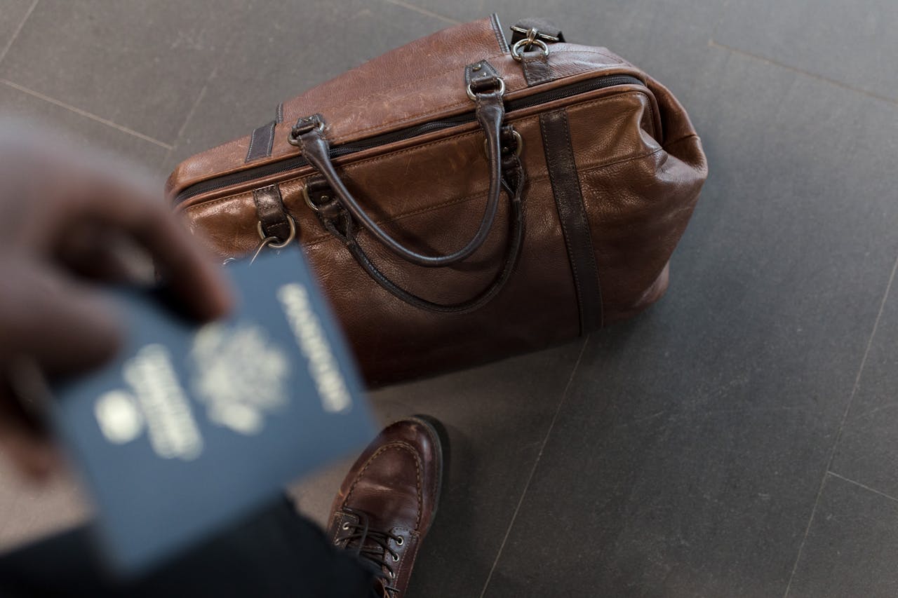 Ein unscharfes Bild einer Person, die einen Reisepass in der Hand hält. Auf dem Boden steht eine braune Ledertasche. Die Person trägt braune Lederschuhe. Die Szene vermittelt den Eindruck einer bevorstehenden Reise.