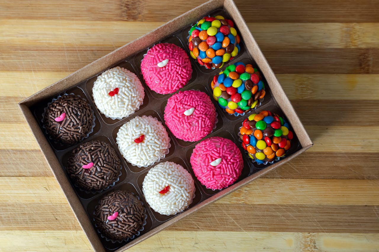 Eine offene Schachtel mit zwölf bunten Pralinen, die in drei Reihen angeordnet sind. Die Pralinen sind mit unterschiedlichen Dekorationen versehen, darunter Schokoladenstreusel, weiße Streusel, rosa Streusel und bunte Schokolinsen. Jede Praline ist in einem kleinen Papierförmchen platziert. Die Schachtel steht auf einem hölzernen Untergrund.