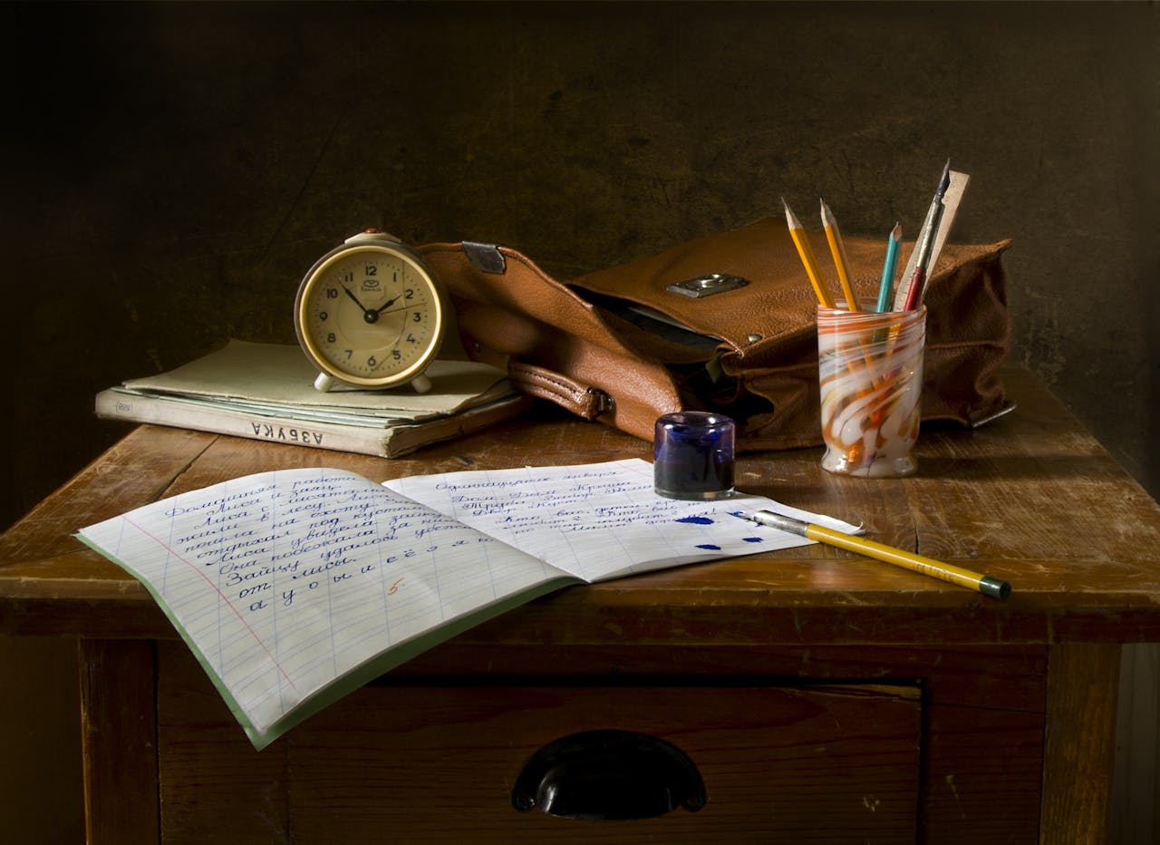 Ein nostalgisches Stillleben auf einem hölzernen Schreibtisch. Darauf liegen ein offenes Notizheft mit handgeschriebenem Text in blauer Tinte, daneben ein gelber Bleistift und ein Tintenfass mit verschütteten Tropfen. Im Hintergrund stehen ein alter Wecker, eine braune Ledertasche und ein Stiftehalter mit Bleistiften und Linealen.