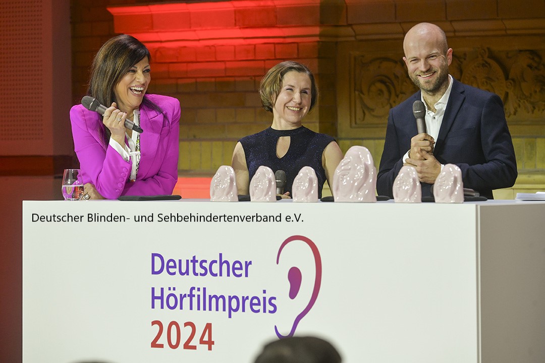 Drei Personen stehen hinter einem Podium bei der Verleihung des Deutschen Hörfilmpreises 2024. Eine Frau im pinken Blazer und zwei weitere Personen, eine Frau und ein Mann, halten Mikrofone und lächeln. Auf dem Podium steht der Schriftzug "Deutscher Hörfilmpreis 2024", daneben das Logo mit einem stilisierten Ohr. Mehrere gläserne Trophäen in Form von Ohren sind auf dem Podium ausgestellt.