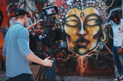 Ein Team dreht ein Video vor einer Wand mit farbenfroher Graffiti-Kunst. Im Vordergrund steht ein Kameramann mit Baseballkappe, der eine professionelle Filmkamera auf einem Stativ bedient. Im Hintergrund gibt ein weiterer Teammitglied Anweisungen und ein Model posiert neben der kunstvoll gestalteten Wand, auf der ein meditatives Gesicht mit geschlossenen Augen dargestellt ist.
