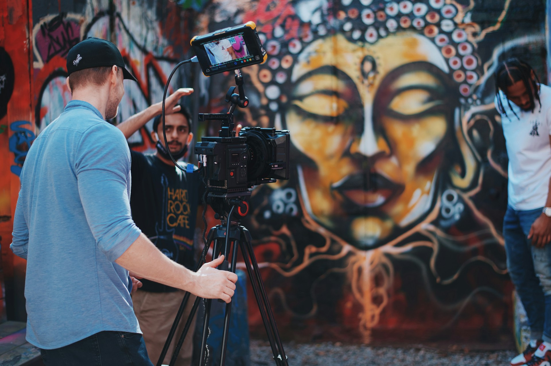 Ein Team dreht ein Video vor einer Wand mit farbenfroher Graffiti-Kunst. Im Vordergrund steht ein Kameramann mit Baseballkappe, der eine professionelle Filmkamera auf einem Stativ bedient. Im Hintergrund gibt ein weiterer Teammitglied Anweisungen und ein Model posiert neben der kunstvoll gestalteten Wand, auf der ein meditatives Gesicht mit geschlossenen Augen dargestellt ist.