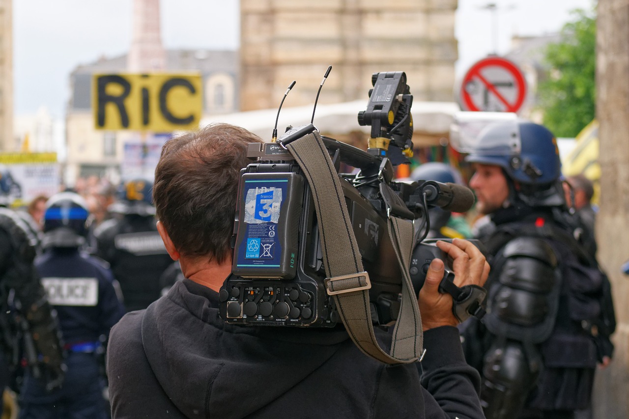 Ein Kameramann nimmt an einer Protestveranstaltung teil und filmt mit einer Schulterkamera das Geschehen. Im Vordergrund sieht man den Kameramann von hinten, während er das Geschehen aufnimmt, das sich vor ihm abspielt. Die Umgebung ist geprägt von einer Menschenmenge und mehreren Polizist in Schutzkleidung, die im Hintergrund sichtbar sind. Ein Plakat mit der Aufschrift »RIC« hebt sich ebenfalls von der Szene ab. Die Szene suggeriert eine Dokumentation von aktuellen politischen Ereignissen, vermutlich im Kontext einer Demonstration.