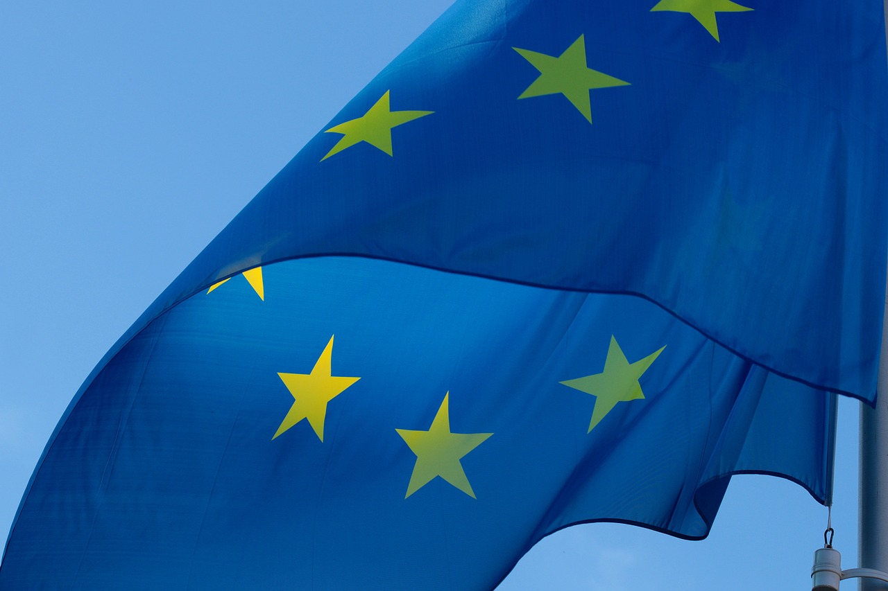 Eine Europaflagge weht im Wind vor einem blauen Himmel. Die Flagge zeigt einen blauen Hintergrund mit gelben Sternen, die im Kreis angeordnet sind und die Einheit der Europäischen Union symbolisieren.