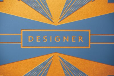 Das Bild zeigt eine stilisierte Grafik mit dem Wort "DESIGNER" in großen Buchstaben. Die Farben Blau und Orange dominieren das Design, das geometrische Muster und feine Details im Art-Deco-Stil verwendet.