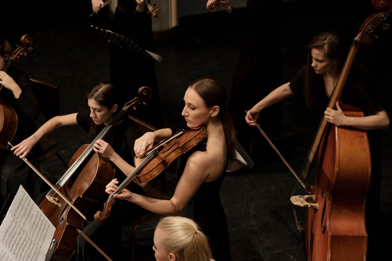 Die Grafik zeigt eine Gruppe von Musikerinnen, die Streichinstrumente spielen. Im Vordergrund sieht man eine Frau mit einer Viola, während um sie herum weitere Musikerinnen auf Cello und Kontrabass konzentriert spielen. Alle tragen formelle, schwarze Kleidung, und die Szene wirkt wie ein klassisches Konzert, das in einem geschlossenen Raum stattfindet.