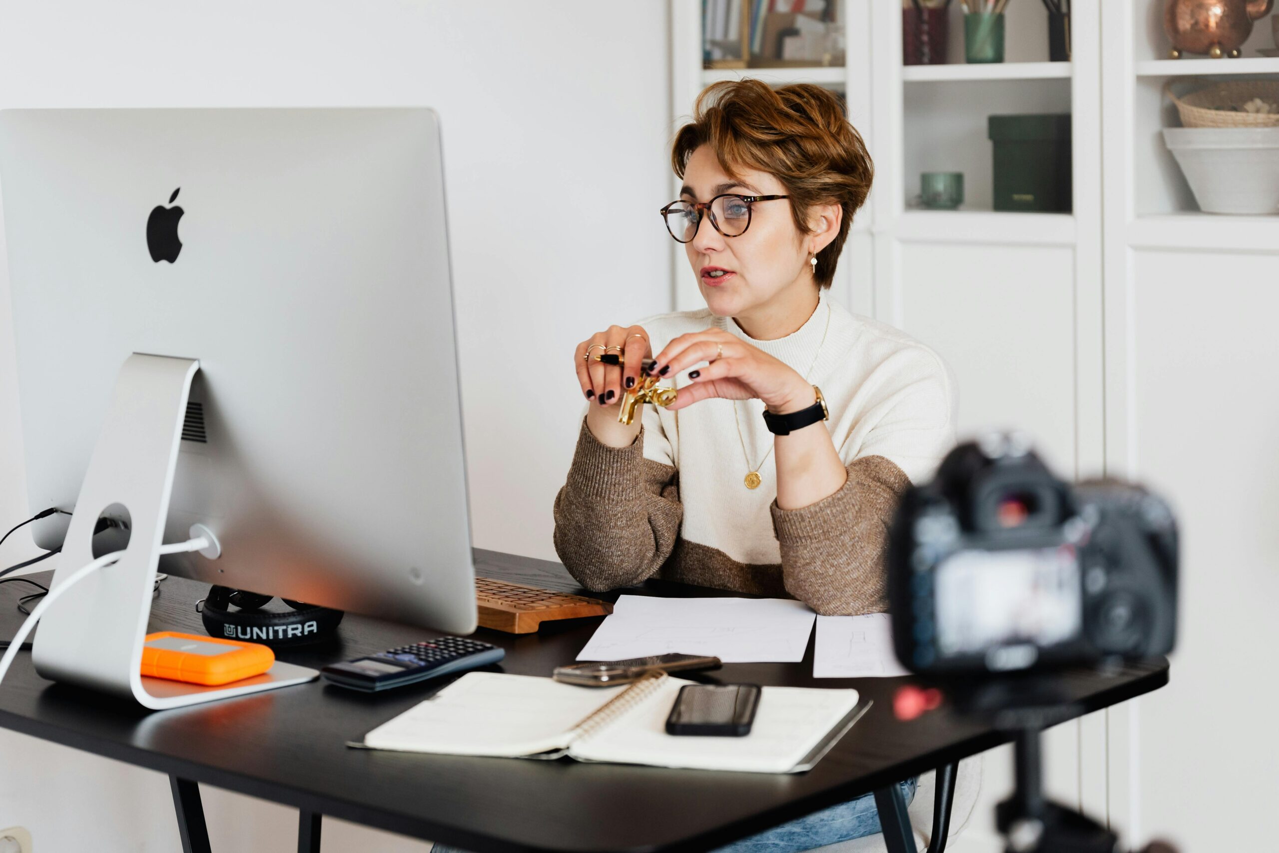 Frau mit Brille und kurzen braunen Haaren sitzt an einem Schreibtisch vor einem großen Computerbildschirm. Sie trägt einen Pullover in Creme und Braun und hält einen goldenen Stift in den Händen. Auf dem Schreibtisch liegen ein Notizbuch, ein Smartphone, eine externe Festplatte, ein Taschenrechner und einige Blätter Papier. Im Vordergrund steht eine Kamera auf einem Stativ, die auf sie gerichtet ist. Im Hintergrund sind Regale mit Büchern und Dekoration zu sehen.