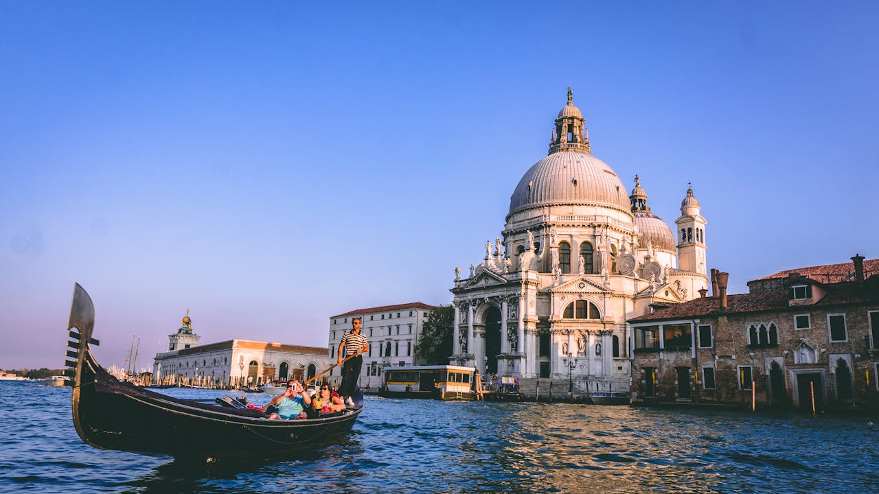 Eine klassische venezianische Gondel mit mehreren Passagier und einem Gondoliere fährt auf dem Wasser vor der imposanten Basilika Santa Maria della Salute in Venedig. Die Abendsonne beleuchtet die historische Architektur und die Wasseroberfläche, während ein wolkenloser, blauer Himmel die Szene umrahmt.