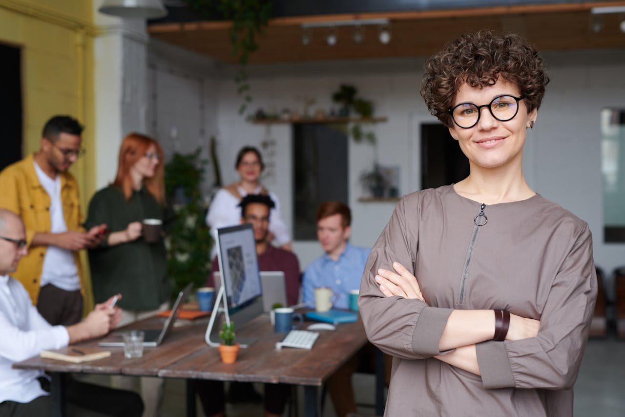 Eine lächelnde Frau mit lockigem Haar und Brille steht im Vordergrund eines modernen Büro-Settings mit verschränkten Armen. Im Hintergrund arbeitet eine Gruppe von Menschen an einem Tisch mit Laptops, Pflanzen und Notizbüchern, während sie miteinander interagieren.