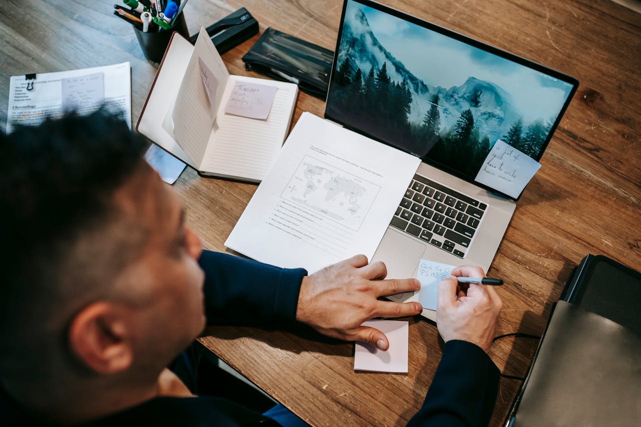 Eine Person arbeitet konzentriert an einem Schreibtisch mit einem Laptop, mehreren Notizbüchern und Dokumenten. Auf dem Laptopbildschirm ist eine Naturaufnahme mit Bergen zu sehen. Die Person macht sich handschriftliche Notizen auf einem Zettel und hat weitere Notizzettel auf dem Laptop befestigt. Der Arbeitsplatz wirkt gut organisiert, mit Schreibutensilien und Papieren.