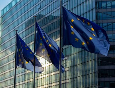 Drei wehende EU-Flaggen mit gelben Sternen auf blauem Hintergrund vor einem modernen Glasgebäude bei Tageslicht.