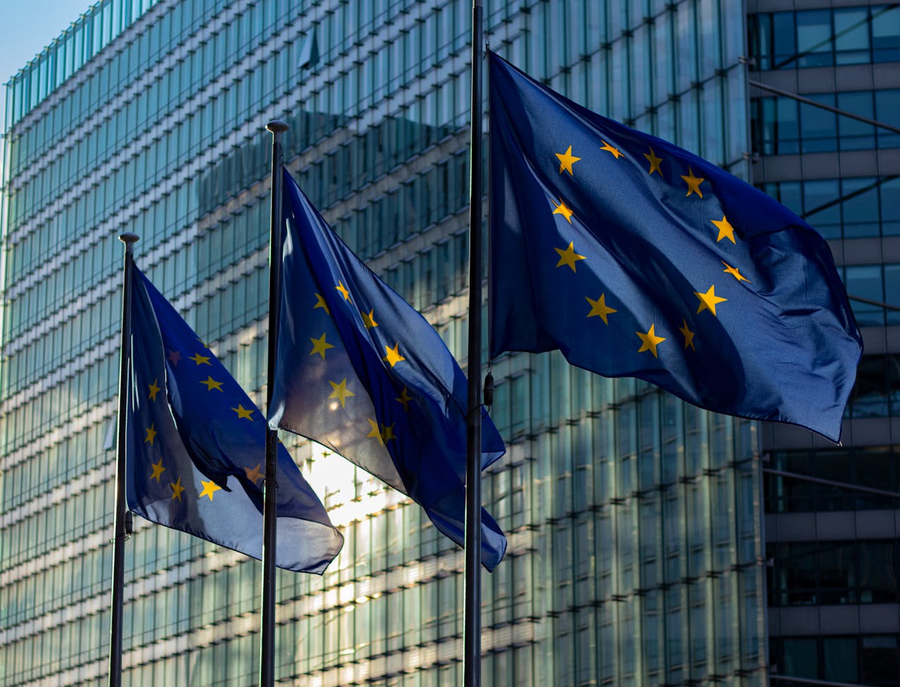 Drei wehende EU-Flaggen mit gelben Sternen auf blauem Hintergrund vor einem modernen Glasgebäude bei Tageslicht.