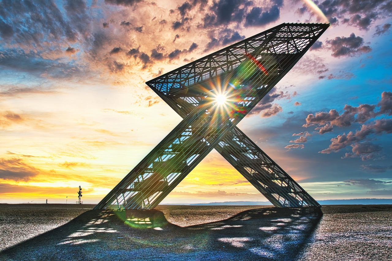 Ein modernes Stahlmonument in X-Form, das Saar-Polygon, steht vor einem beeindruckenden Sonnenuntergang, wobei Sonnenstrahlen durch die Struktur brechen. Im Hintergrund sind Wolken am farbenfrohen Himmel und eine weite, offene Landschaft zu sehen.