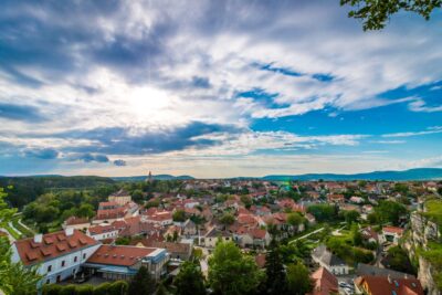 Panorama einer kleinen Stadt mit roten Ziegeldächern, umgeben von grünen Wäldern und Hügeln unter einem blauen Himmel mit weißen Wolken. Im Hintergrund sind eine Kirche mit Turm und eine weite, malerische Landschaft zu sehen.