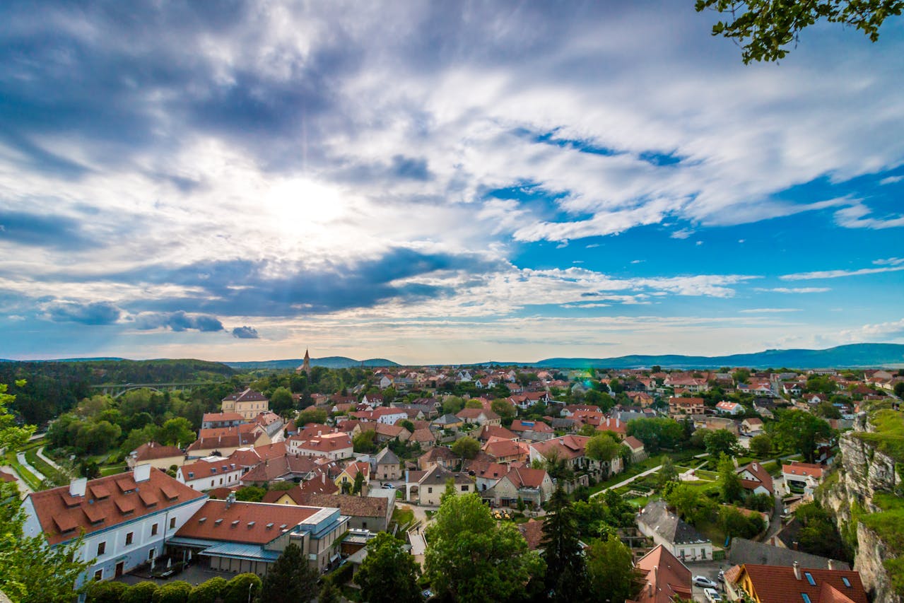 Panorama einer kleinen Stadt mit roten Ziegeldächern, umgeben von grünen Wäldern und Hügeln unter einem blauen Himmel mit weißen Wolken. Im Hintergrund sind eine Kirche mit Turm und eine weite, malerische Landschaft zu sehen.