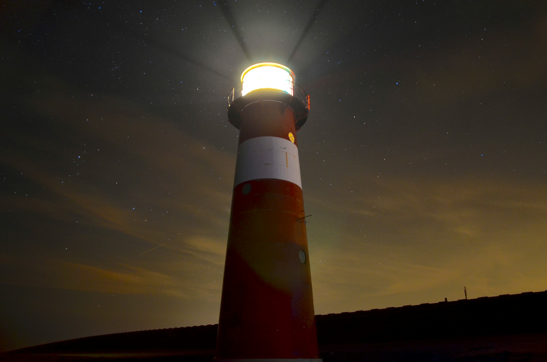 Ein rot-weiß gestreifter Leuchtturm leuchtet in die Dunkelheit und strahlt Lichtstrahlen in den Nachthimmel. Sterne sind im Hintergrund sichtbar, und der Himmel ist leicht bewölkt, was eine mystische Atmosphäre erzeugt.