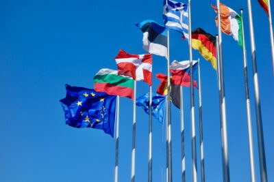 Mehrere Flaggen europäischer Länder und der Europäischen Union wehen im Wind vor einem klaren, blauen Himmel. Die Flaggen sind auf hohen Masten gehisst und symbolisieren die Einheit und Zusammenarbeit der europäischen Nationen.