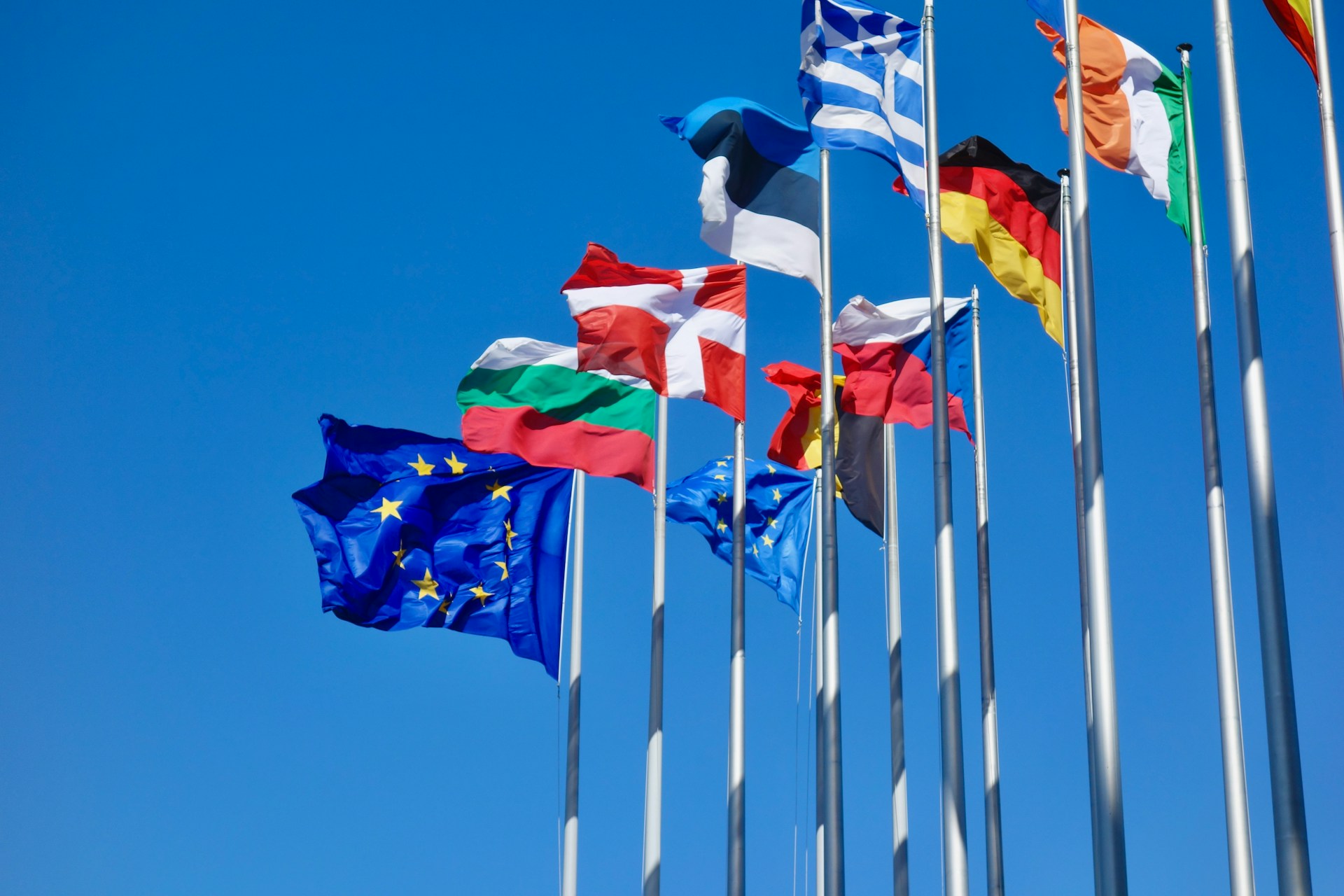 Mehrere Flaggen europäischer Länder und der Europäischen Union wehen im Wind vor einem klaren, blauen Himmel. Die Flaggen sind auf hohen Masten gehisst und symbolisieren die Einheit und Zusammenarbeit der europäischen Nationen.