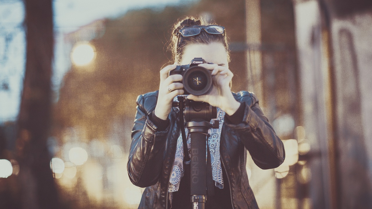 Eine Fotografin steht hinter ihrer Kamera und richtet den Fokus auf ein Motiv. Sie trägt eine Lederjacke und hält die Kamera mit beiden Händen. Im Hintergrund sind unscharfe Lichter und eine städtische Umgebung bei Dämmerung zu sehen. Die Szene vermittelt Kreativität und Leidenschaft für die Fotografie.