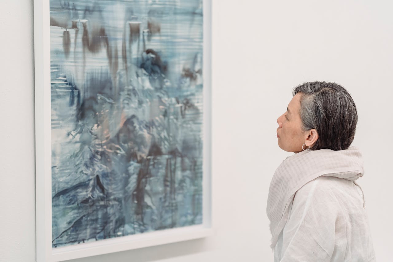 Eine Frau mit grauem, kurzem Haar betrachtet ein abstraktes Gemälde in blauen und grauen Farbtönen in einer Kunstgalerie. Sie trägt einen hellen Schal und eine weiße, lockere Bluse. Das Bild hat eine ruhige und nachdenkliche Atmosphäre.