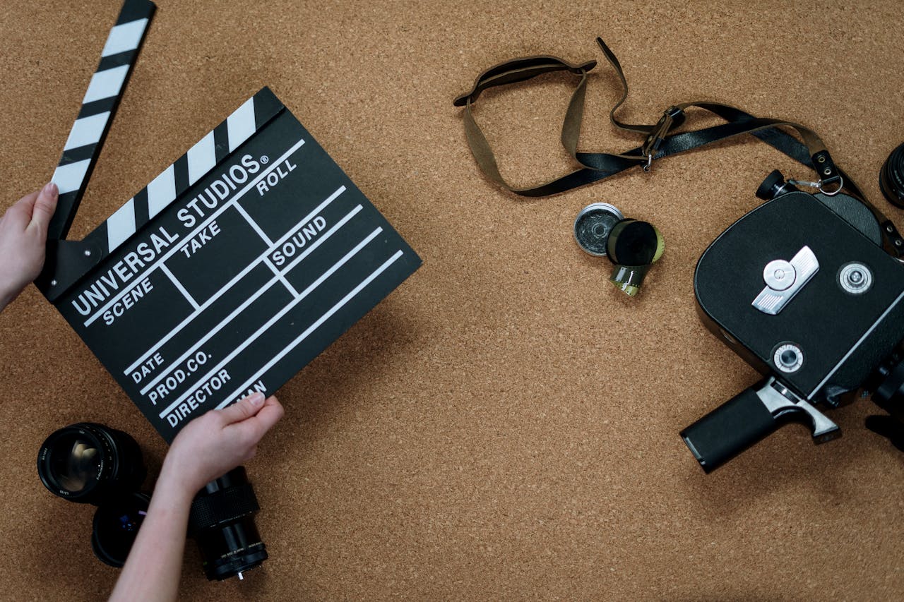 Eine Person hält eine schwarze Filmklappe mit der Aufschrift „Universal Studios“. Auf dem Tisch liegen eine alte Filmkamera, Linsen und Filmzubehör verstreut. Der Hintergrund besteht aus einer hellbraunen, strukturierten Korkplatte.