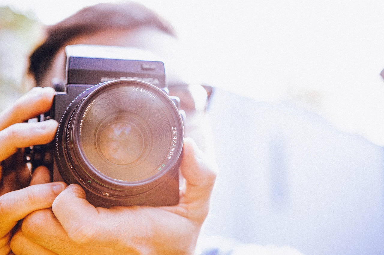 Ein:e Fotograf:in hält eine Kamera im Querformat, der Fokus liegt auf dem Objektiv. Im Hintergrund ist eine helle, verschwommene Umgebung zu sehen.