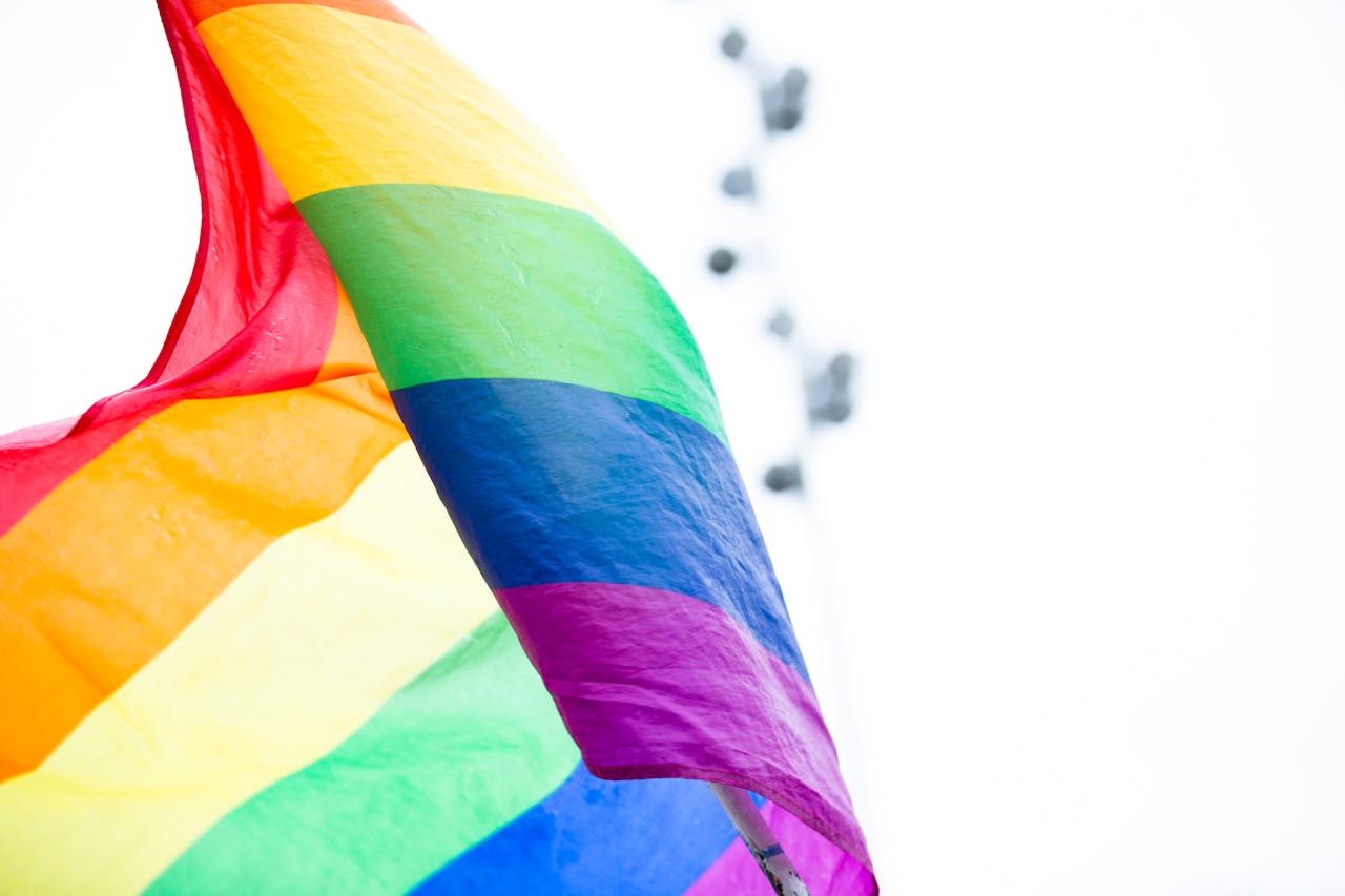 Eine Regenbogenflagge, Symbol der LGBTQIA+-Gemeinschaft, weht bei Tageslicht vor einem weißen, unscharfen Hintergrund.