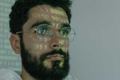 Ein Mann mit Brille und Bart blickt konzentriert nach vorne. Grüner Binärcode wird auf sein Gesicht projiziert. Der Hintergrund ist neutral und hell, wodurch der Fokus auf dem projizierten Code und seinem Gesicht liegt.