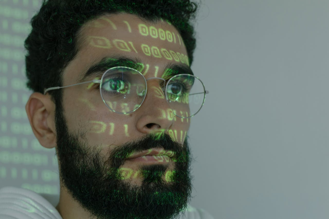 Ein Mann mit Brille und Bart blickt konzentriert nach vorne. Grüner Binärcode wird auf sein Gesicht projiziert. Der Hintergrund ist neutral und hell, wodurch der Fokus auf dem projizierten Code und seinem Gesicht liegt.