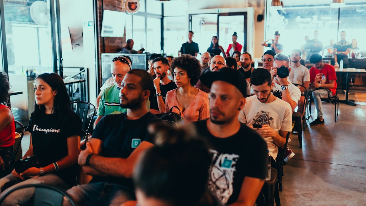 Eine Gruppe von Menschen sitzt aufmerksam in einem Veranstaltungsraum mit modernen, hellen Elementen. Im Vordergrund sind diverse Personen unterschiedlichen Geschlechts und Alters zu sehen, einige mit Kameras oder Smartphones. Die Atmosphäre wirkt konzentriert, als ob sie einer Präsentation oder einem Vortrag lauschen. Im Hintergrund sind weitere Teilnehmer sowie Fenster und Lichteinfall zu erkennen.