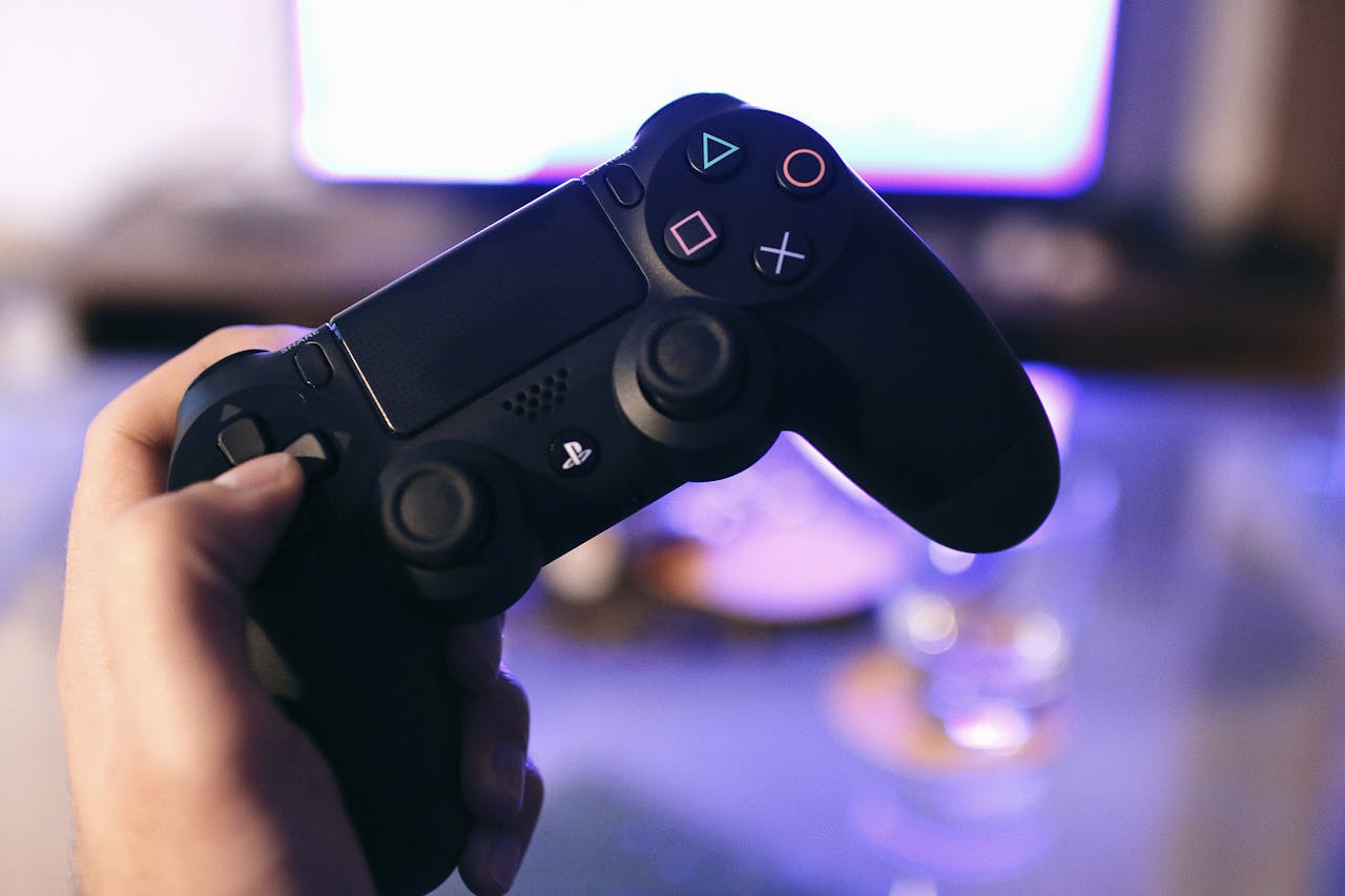 Eine Hand hält einen schwarzen Gaming-Controller, der deutlich die farbigen Tasten und Analogsticks zeigt. Im Hintergrund ist ein unscharfer Bildschirm mit violettem Licht zu erkennen, was eine entspannte Gaming-Atmosphäre andeutet.