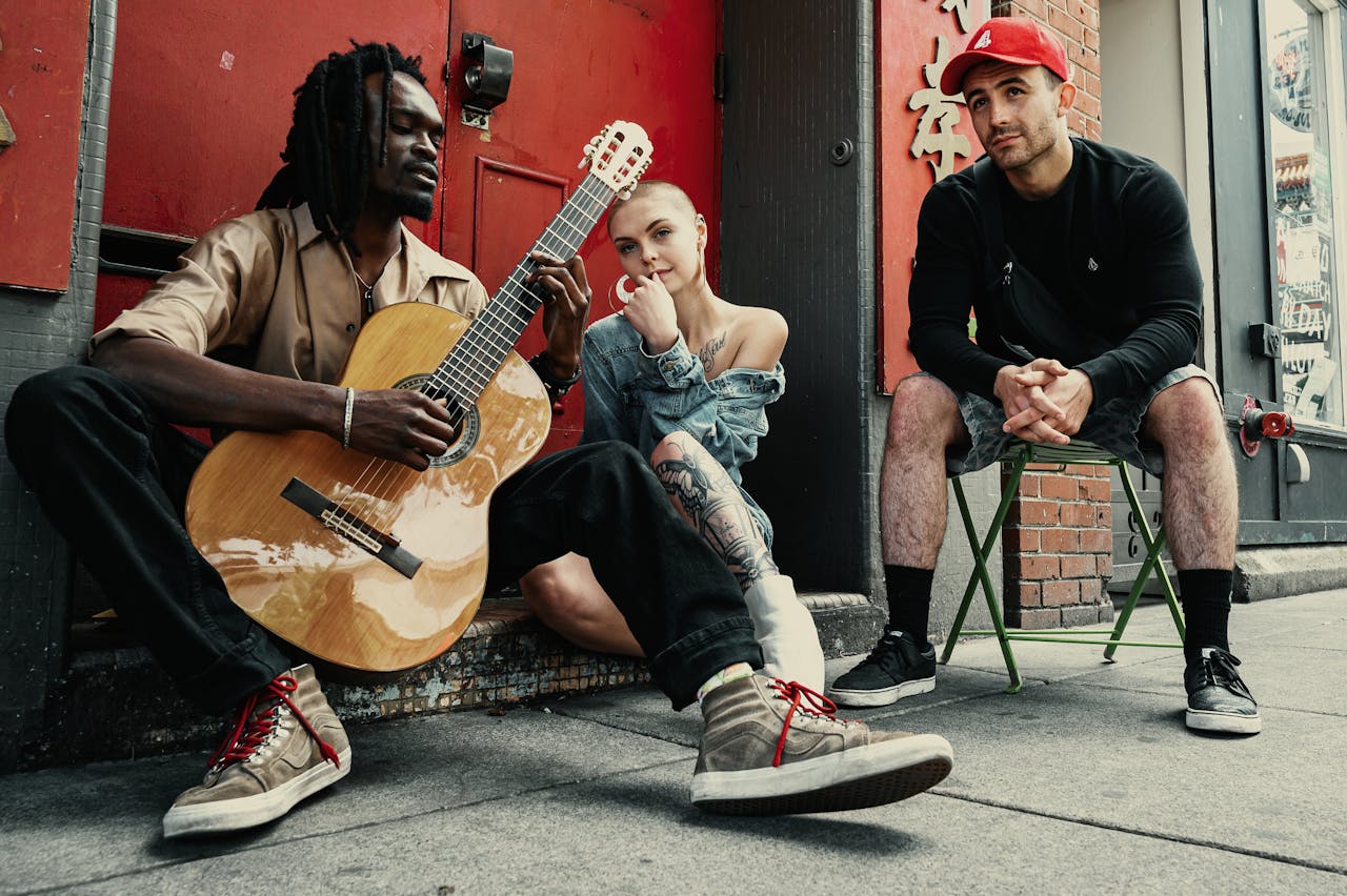 Drei junge Menschen sitzen vor einer roten Tür auf einer Straße. Der Mann links, mit dunklen Dreadlocks und einem beigen Hemd, spielt eine akustische Gitarre. Die Frau in der Mitte, mit rasiertem Kopf, großen Ohrringen und einem Jeans-Oberteil, sitzt lässig auf dem Boden und schaut nachdenklich. Der Mann rechts, in einem schwarzen Pullover, Camouflage-Shorts und einer roten Baseballkappe, sitzt auf einem grünen Klappstuhl und beobachtet die Szene entspannt. Die Atmosphäre wirkt urban und kreativ.