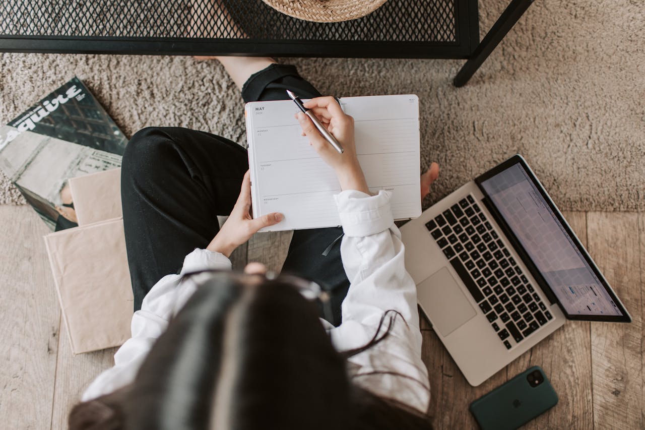 Eine Frau sitzt auf dem Boden neben einem Couchtisch und arbeitet. Sie hält einen Stift in der Hand und schreibt in einen offenen Planer. Neben ihr liegt ein aufgeklappter Laptop, ein Smartphone und einige Bücher. Der Raum hat einen Holzboden mit einem Teppich, und eine Zeitschrift ist ebenfalls sichtbar.