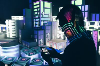 Silhouette einer Person mit Brille, die ein Smartphone hält, während bunte digitale Projektionen von futuristischen Gebäuden den Hintergrund erleuchten.