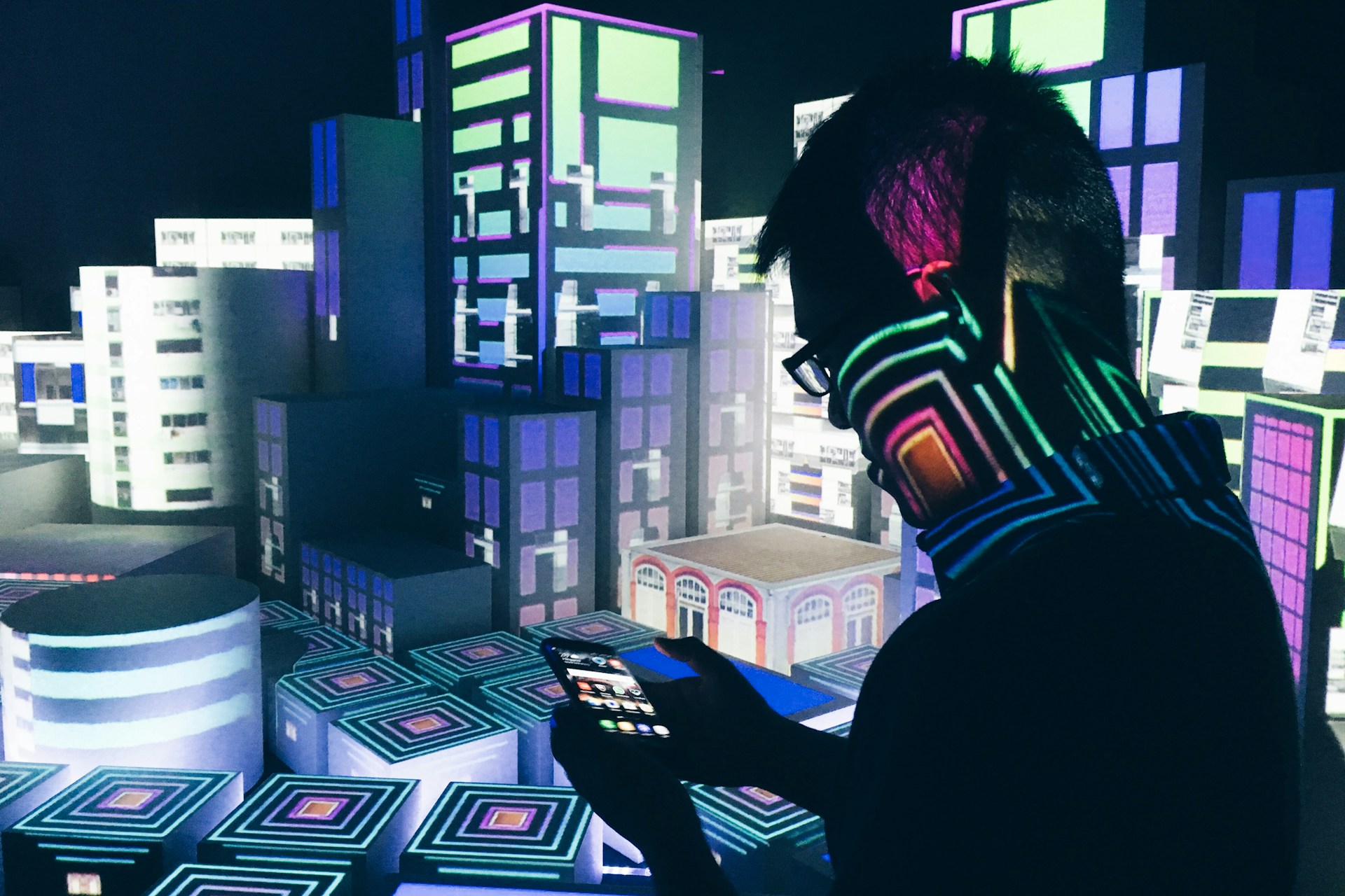 Silhouette einer Person mit Brille, die ein Smartphone hält, während bunte digitale Projektionen von futuristischen Gebäuden den Hintergrund erleuchten.